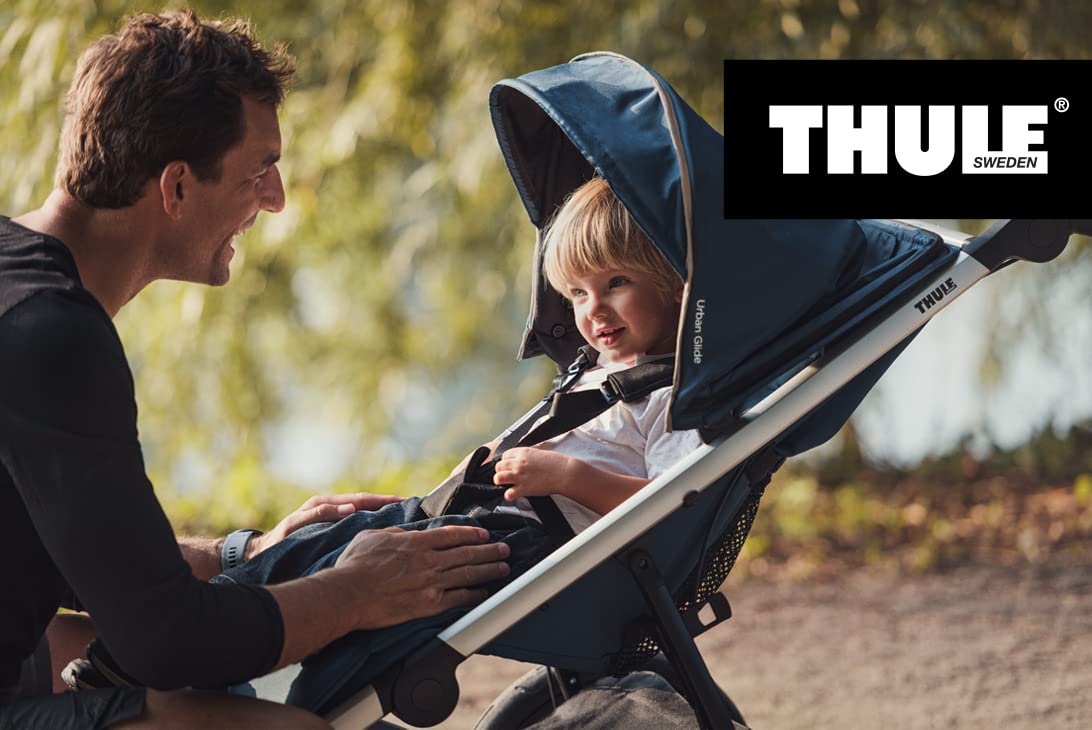 Parent kneels beside child in Thule stroller during outdoor walk, with sunlit foliage in background