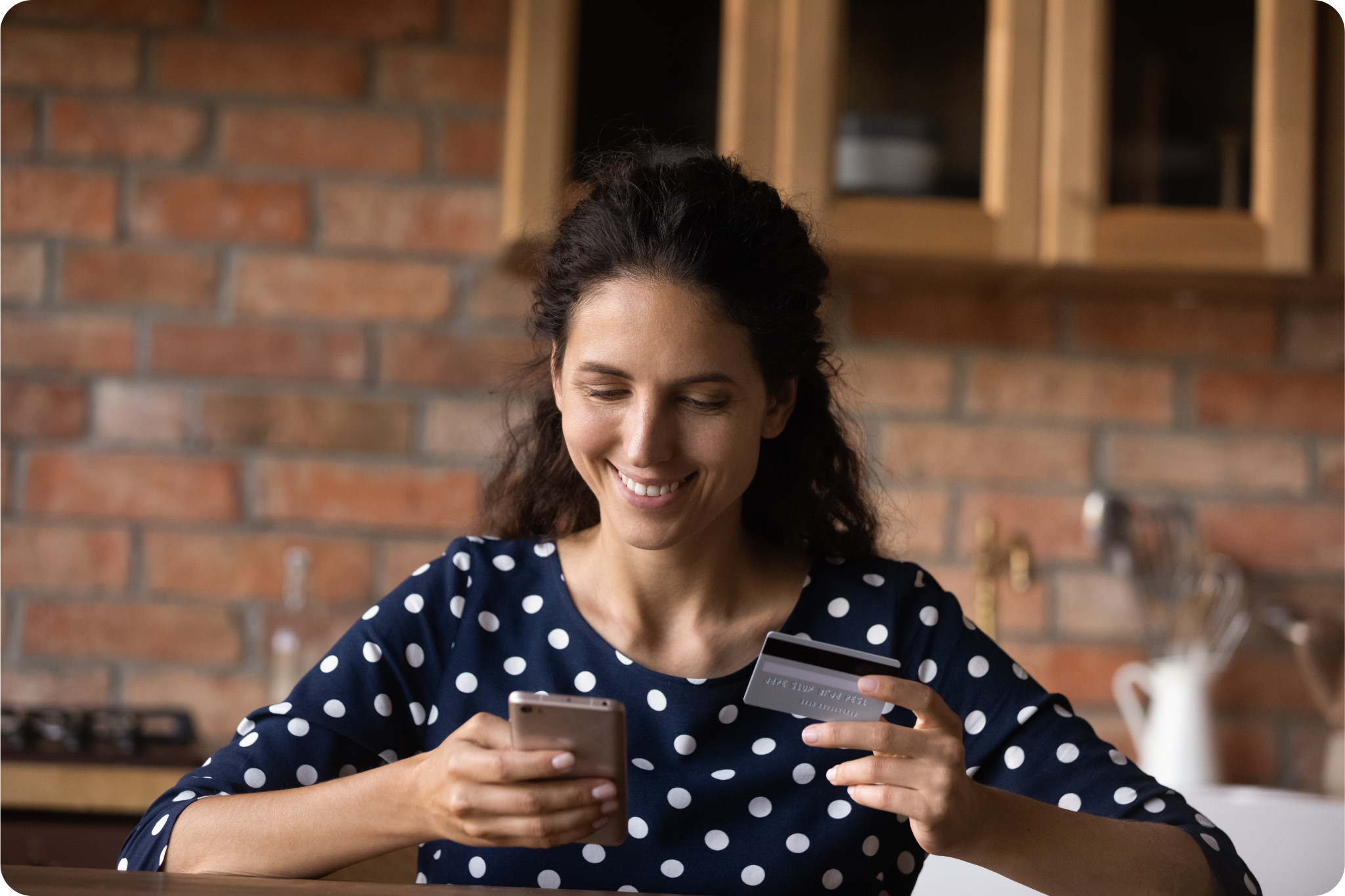 Mulher segurando celular e cartão de crédito em uma cozinha com parede de tijolos