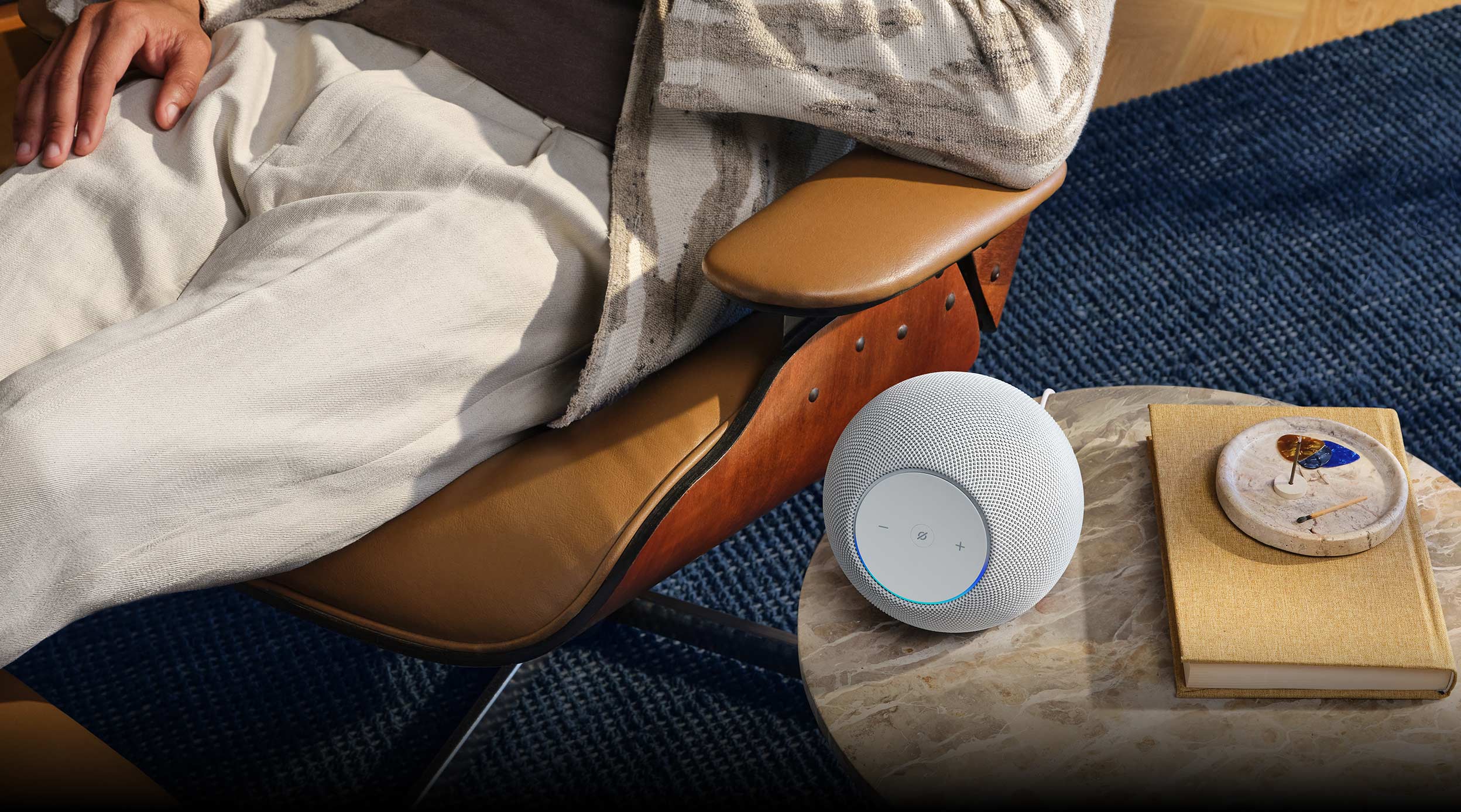 Person relaxing in lounge chair with an Echo Dot Max speaker on a small round table nearby, alongside a book and decorative item on a blue area rug.