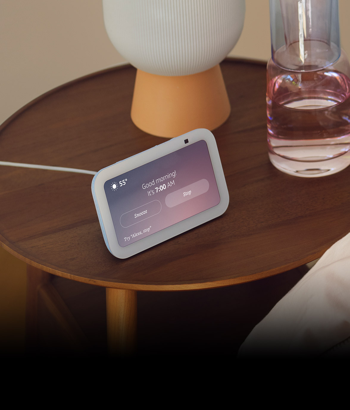 Echo Show 5 on a wooden nightstand beside a lamp and water glass, a person's arm rests in bed with blue sleeves and pink bedding.