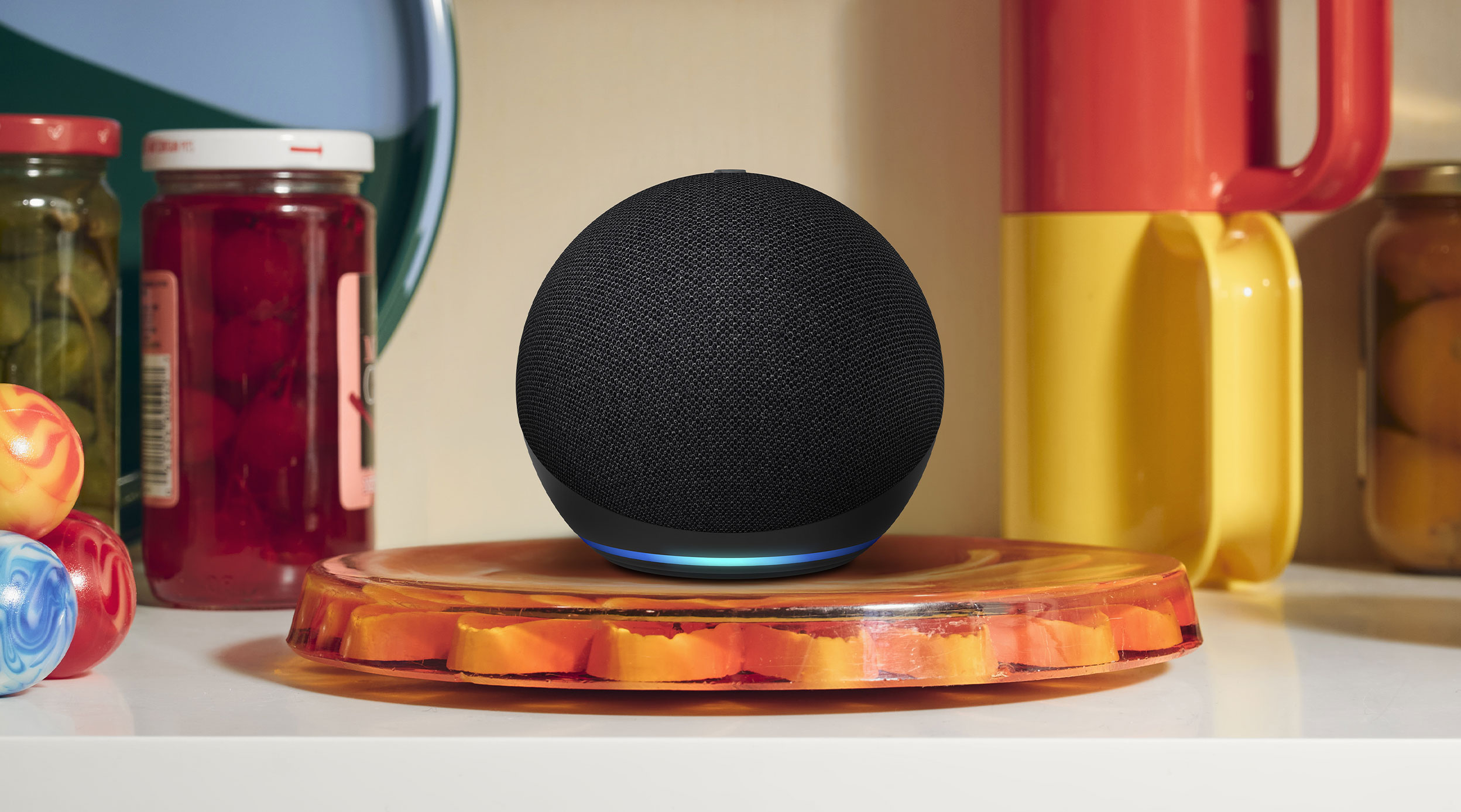 Echo Dot 4-inch spherical speaker with flat bottom and blue light ring sits on amber cutting board in kitchen with jars and colorful mugs.