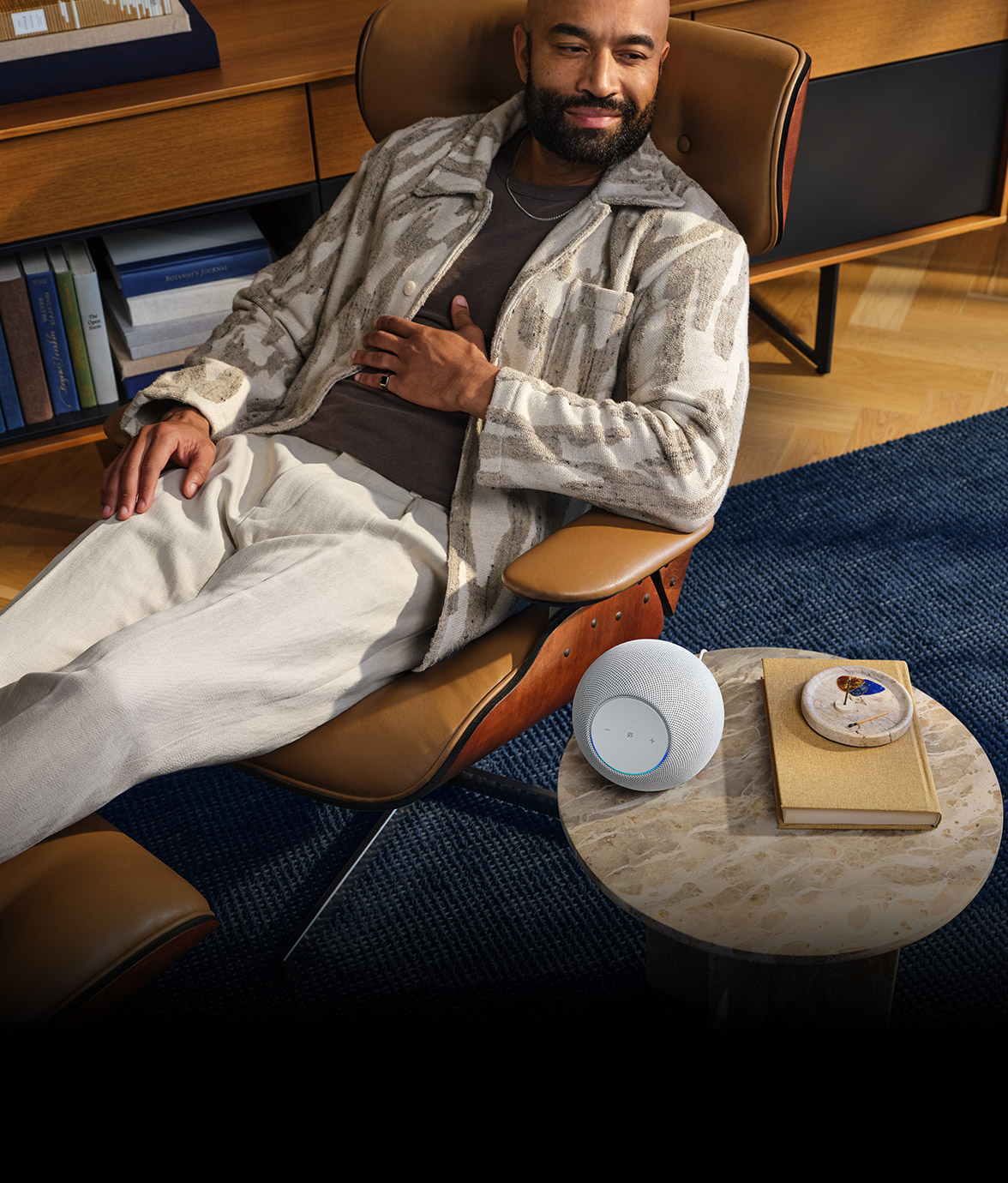 Person relaxing in lounge chair with an Echo Dot Max speaker on a small round table nearby, alongside a book and decorative item on a blue area rug.