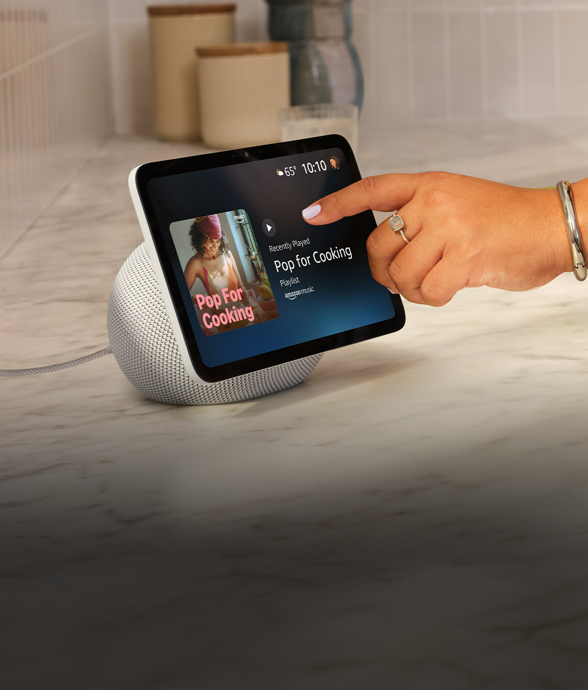 Echo Show 8 smart display with white base. A hand with blue nail polish touches the screen showing a "Pop for Cooking" playlist. Temperature: 65°, time: 10:10. Device sits on marble countertop with storage containers in background.