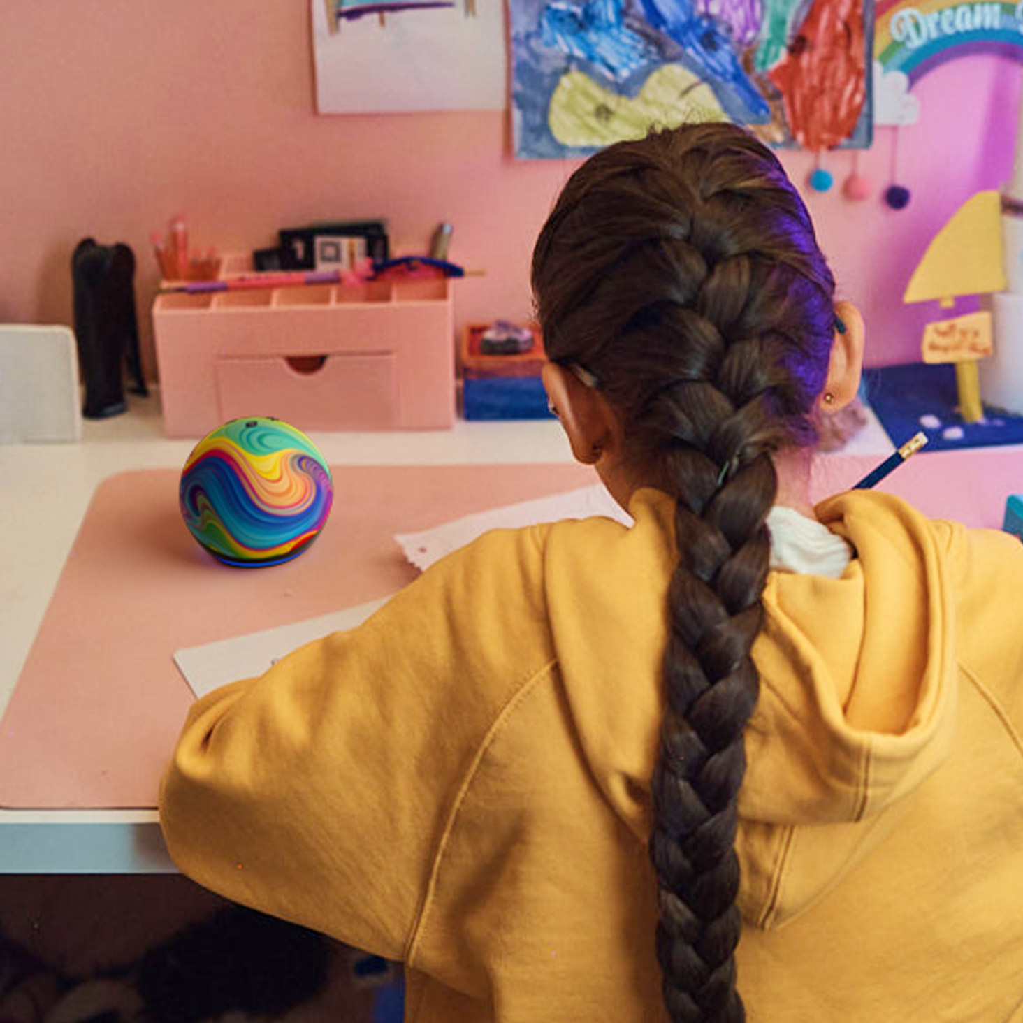 Young girl with braided hair wearing a yellow hoodie sits at a desk with colorful rainbow ball in a child's bedroom with pink walls and children's artwork displayed.