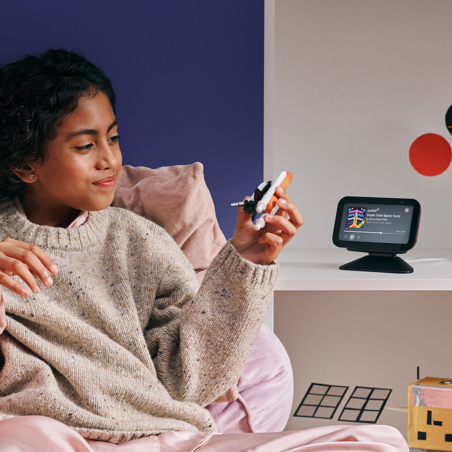 Young girl holding a toy rocket while looking at an Echo Dot 5th Gen displaying space facts on its screen. The smart speaker is positioned on a white shelf against a purple background.