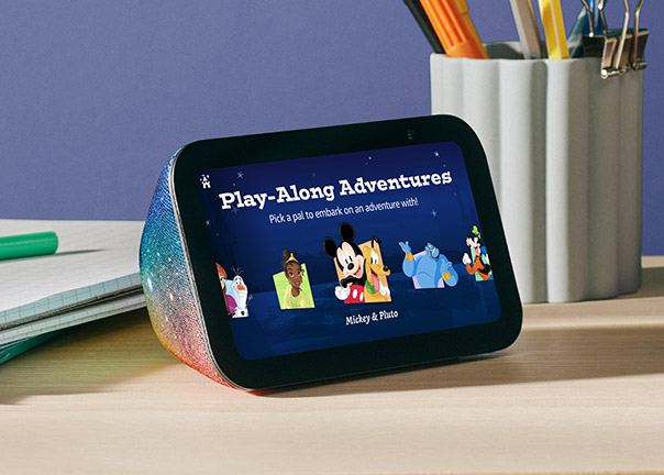 Echo Dot 5th Gen on a wooden desk displaying Disney-themed "Play-Along Adventures" screen with Mickey and other characters. Text reads: "Pick a pal to embark on an adventure with."
