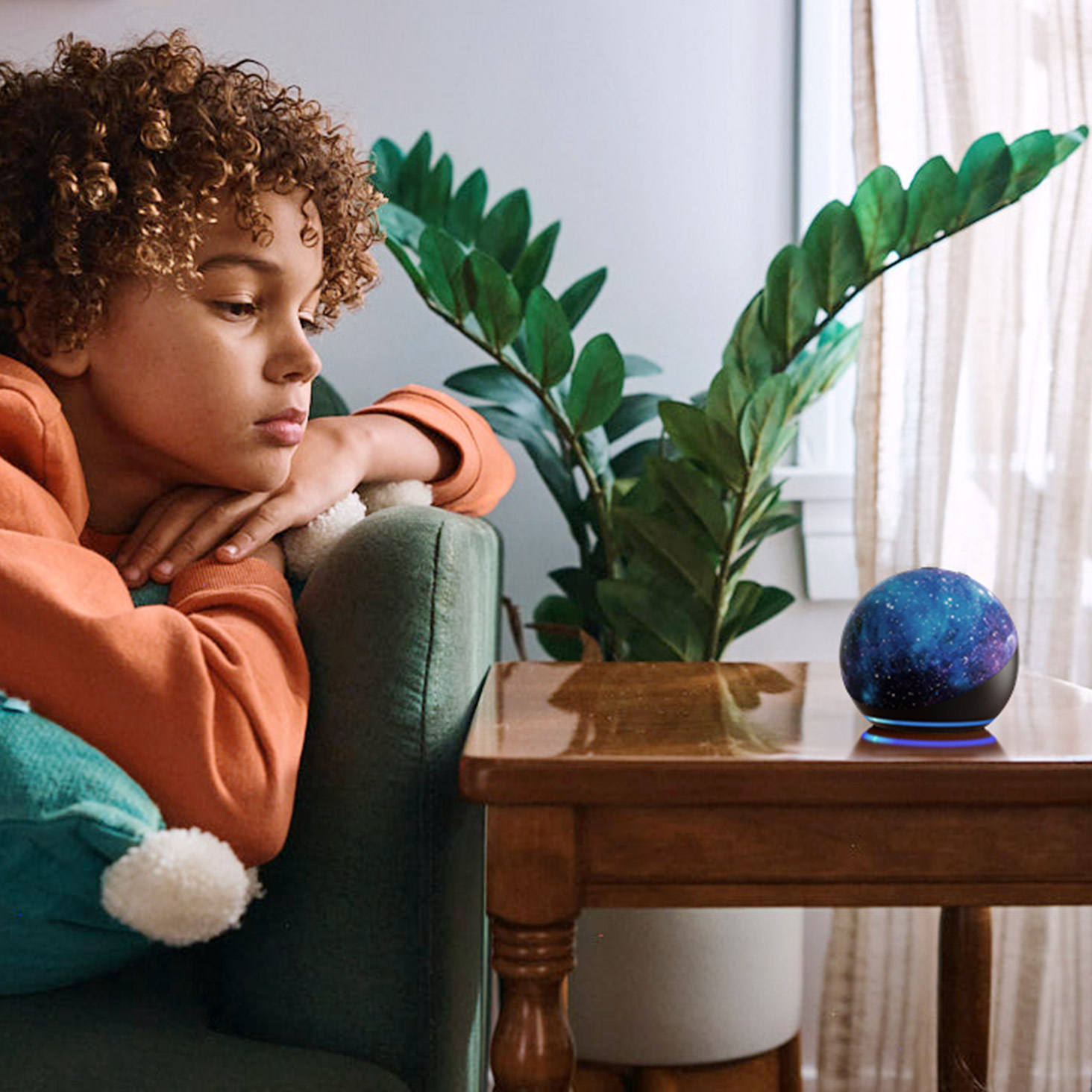 Child with curly hair in orange hoodie resting on green couch looking at Echo Dot with cosmic blue design on wooden side table next to a potted plant.