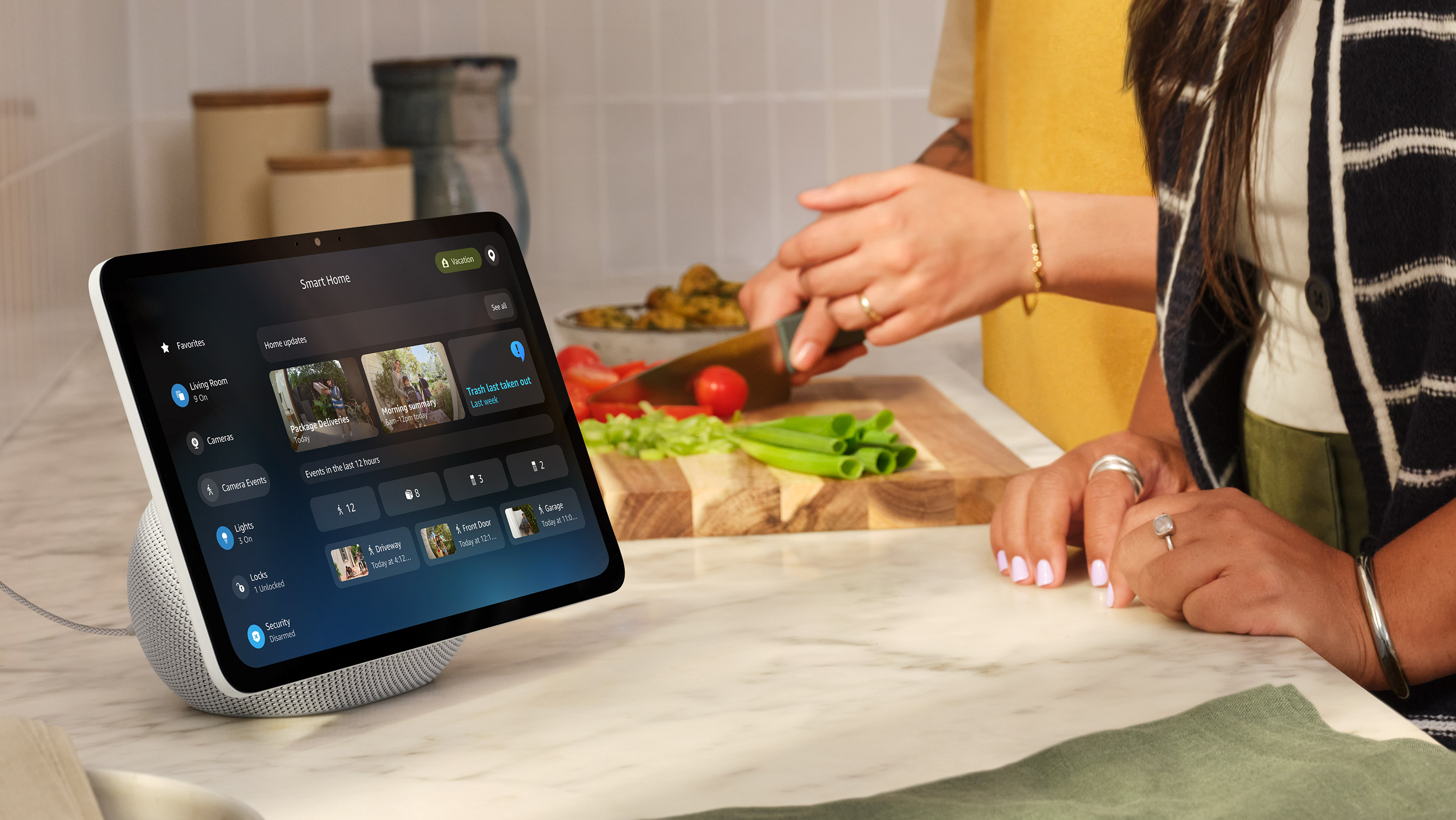 Echo Show displaying Smart Home interface on a kitchen counter while someone chops vegetables on a cutting board. Screen shows Living Room, Cameras, Lights, and Security controls.