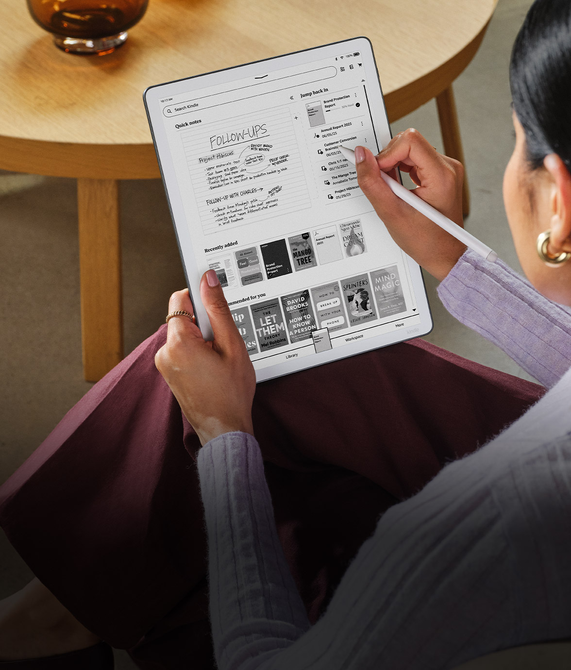 A person sitting with a Kindle Scribe on their lap, using the stylus to navigate notes and documents on the home screen, which shows handwritten follow-up notes and a library of books.