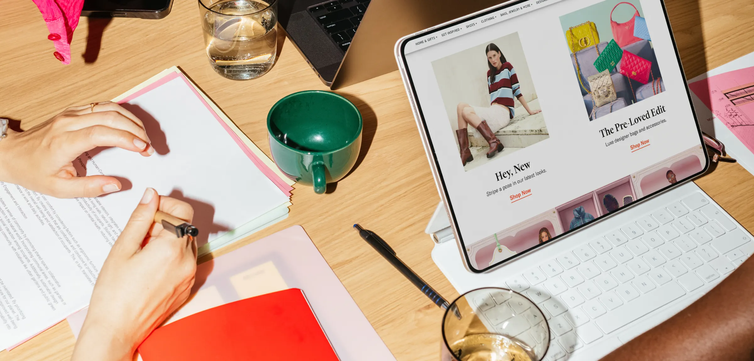 A person writes notes on paper beside a tablet displaying an online Amazon fashion store 