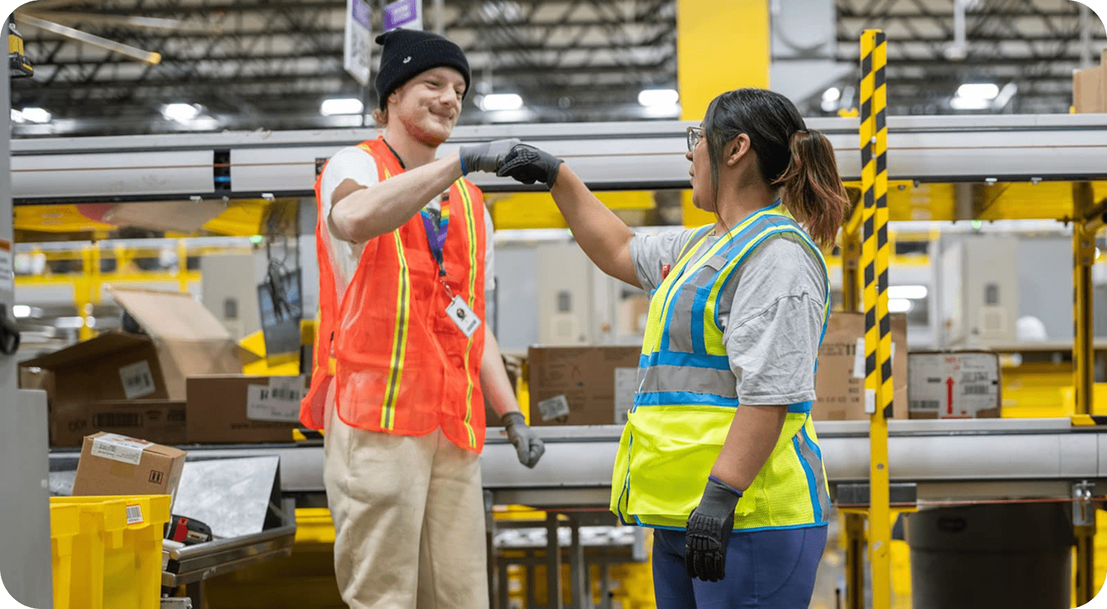 A male associate and female associate doing a fist bump while inside the warehouse. They are wearing safety vests and safety gloves.