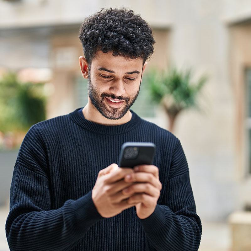 Photo of a male standing outside while smiling and typing into a smartphone. 