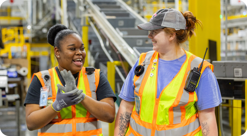 Two Amazon warehouse associates in safety vests smiling and talking on the warehouse floor