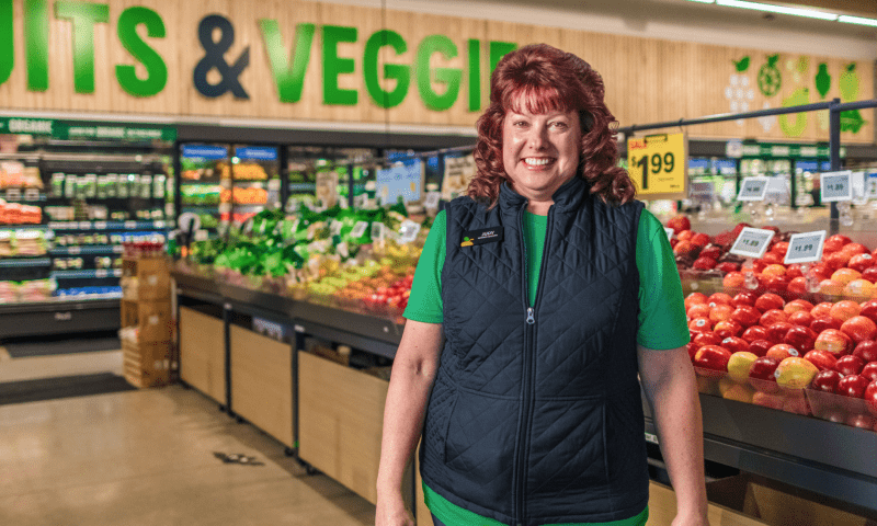 Asociada de Amazon Fresh vistiendo un chaleco de Amazon Fresh. Está sonriendo mientras está de pie en la sección de vegetales y frutas en un supermercado.