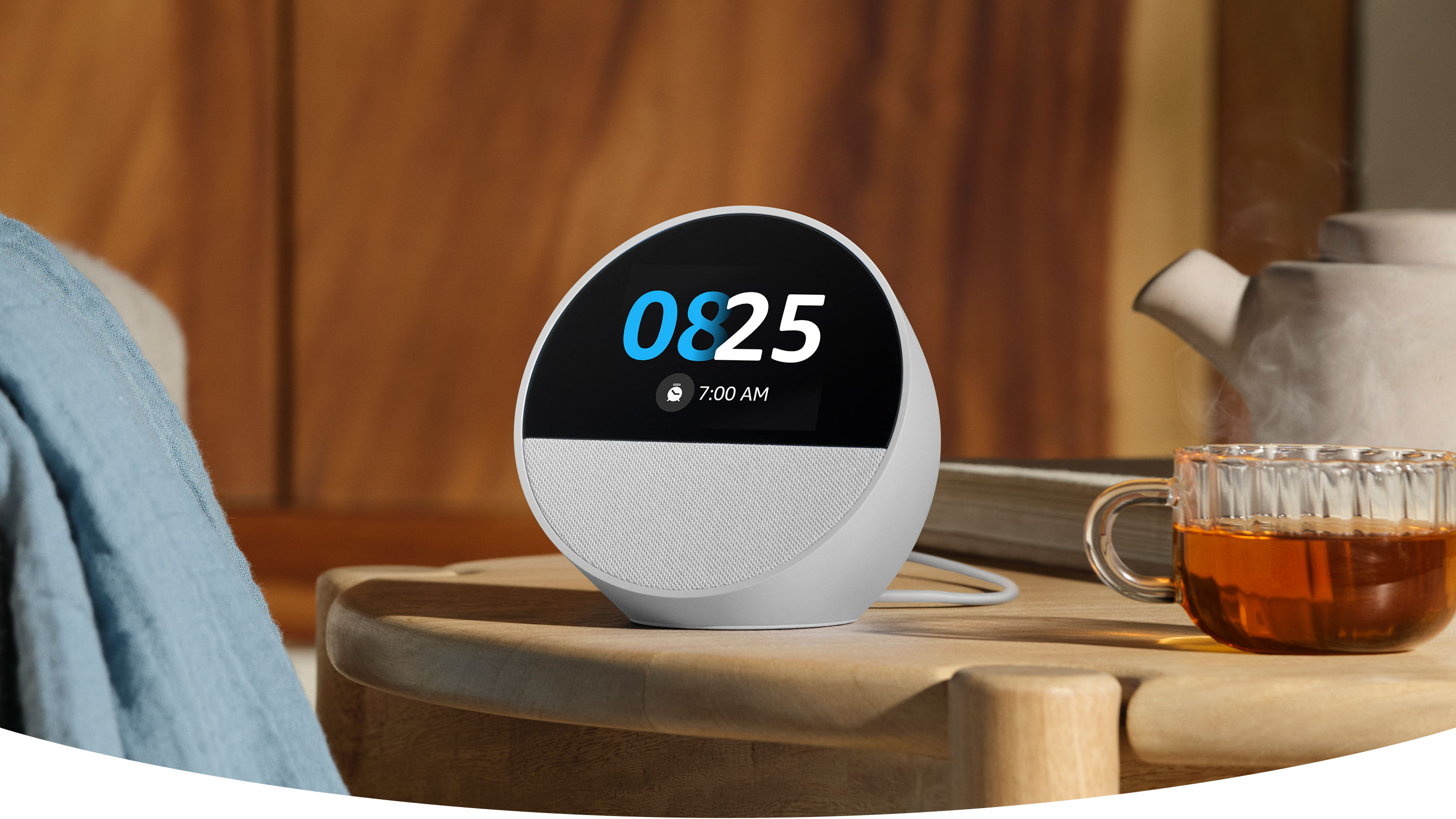 Echo Spot, a semi-spherical smart speaker with a 2.8-inch touch display and a mesh speaker below, on a night stand. The display shows the time, an alarm clock icon and an alarm for 7AM.