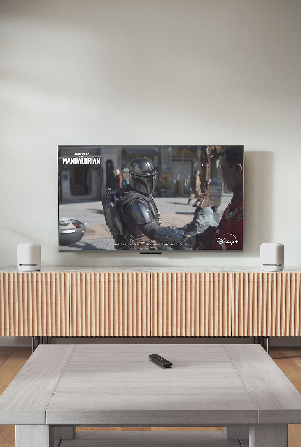 MWJjNDU1MDgt. CB578323195 Ein HDTV in einem modernen Wohnzimmer zeigt „The Mandalorian“. Auf dem Tisch darunter zwei Echo Studio: zylinderförmige Mesh-Lautsprecher mit flacher Ober- und Unterseite sowie vier erhöhten Tasten oben.