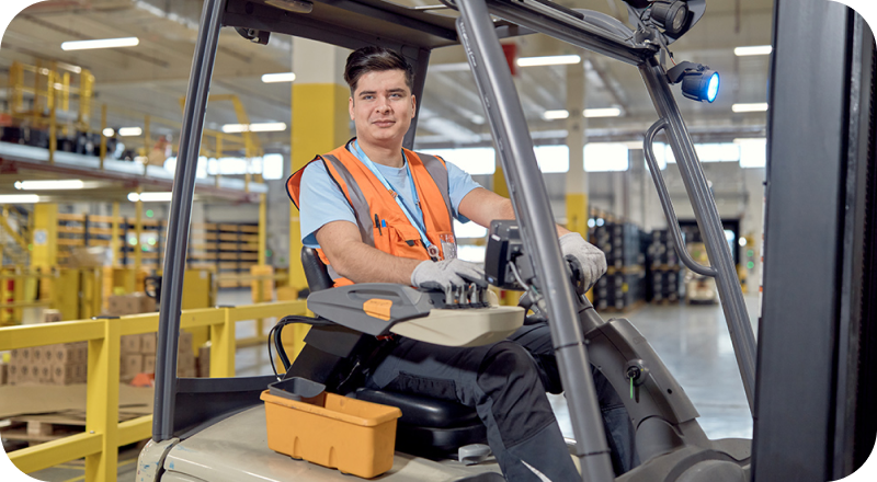Male associate in the warehouse operating a Powered Industrial Truck and smiling while wearing a safety vest.