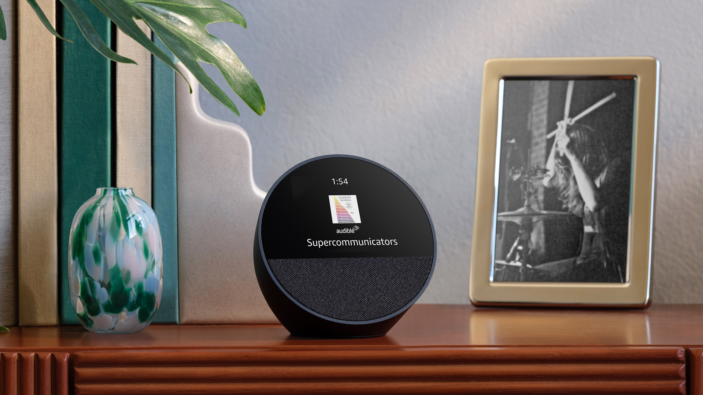 Echo Spot is on an entry table next to a framed photo and a set of books. The 2.8-inch display shows the time and the cover of an Audible book, "Supercommunicators."