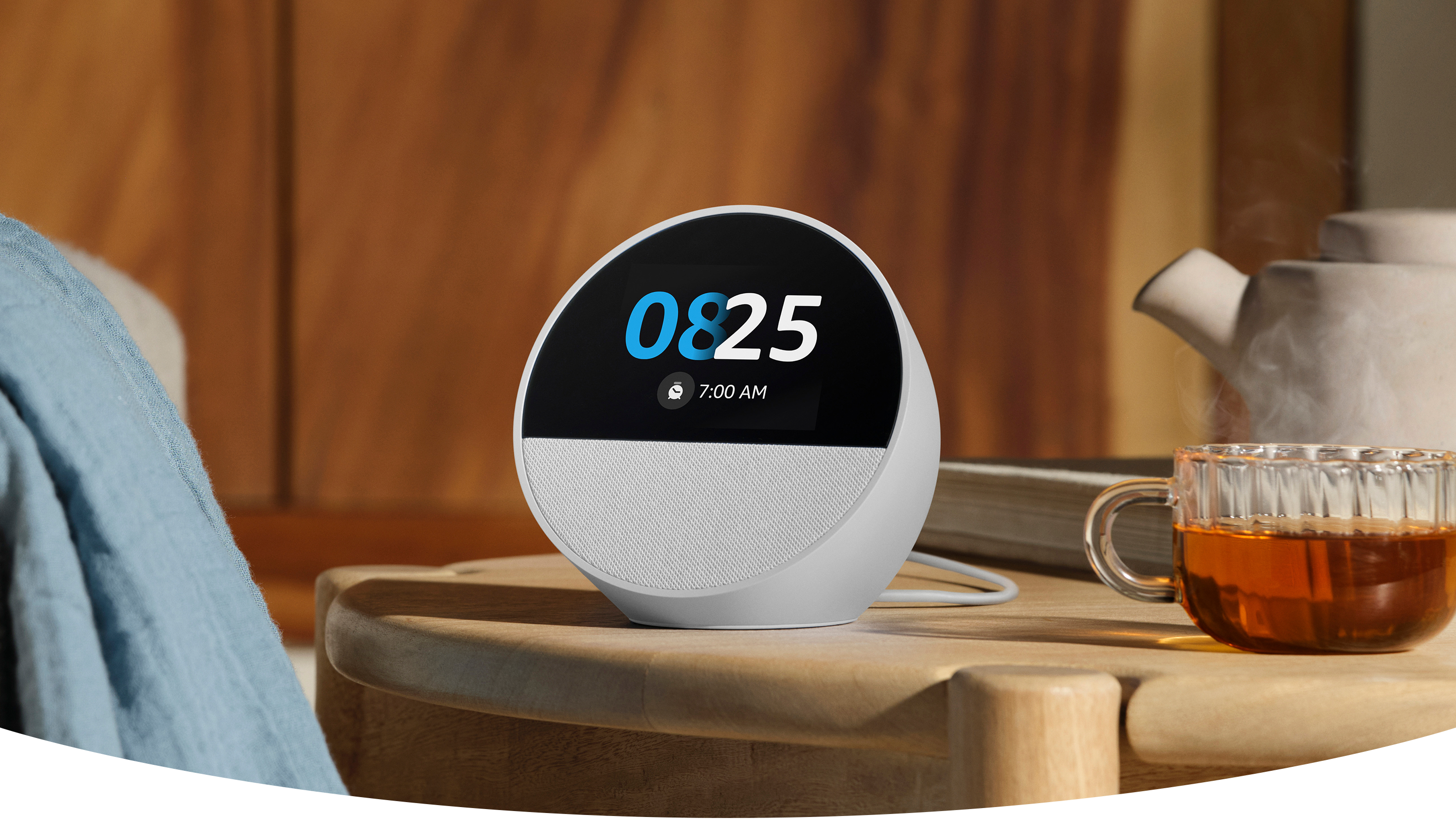 Echo Spot, a semi-spherical smart speaker with a 2.8-inch touch display and a mesh speaker below, on a night stand. The display shows the time, date, weather, and a full-sun weather icon.