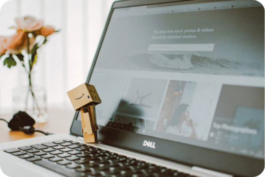 Boneco de papelão apoiado sobre o teclado de um notebook Dell, com a tela exibindo um site. Ao fundo, há um vaso com flores desfocado