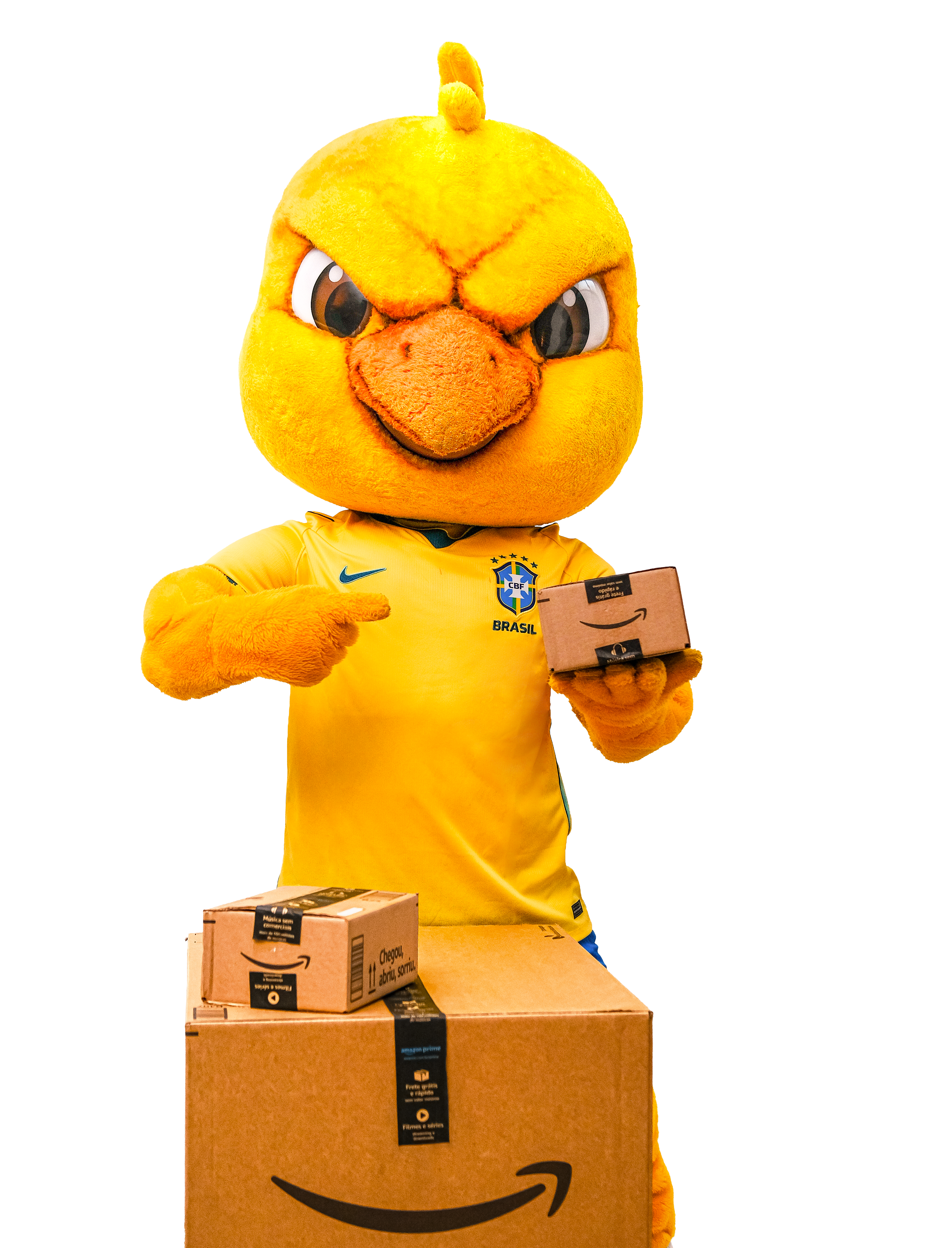 Mascote em formato de pássaro amarelo, com bico laranja e olhos expressivos, vestindo a camisa oficial da Seleção Brasileira de Futebol com o escudo da CBF. O mascote está em pé e segura uma caixa de papelão com o logo da Amazon.