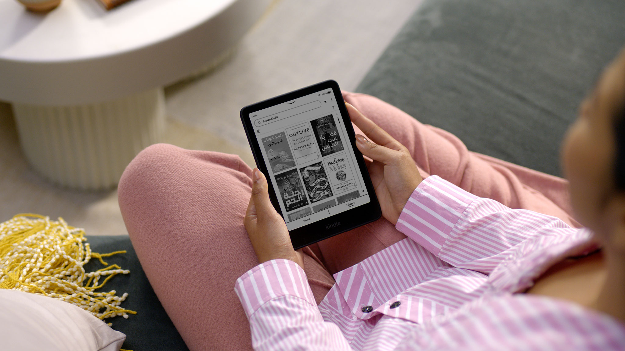 Over the shoulder view of a woman using her Kindle Paperwhite Signature Edition in a living room. She opens a book in the Kindle Library, flips through a few pages, and returns to the Kindle Library page.
