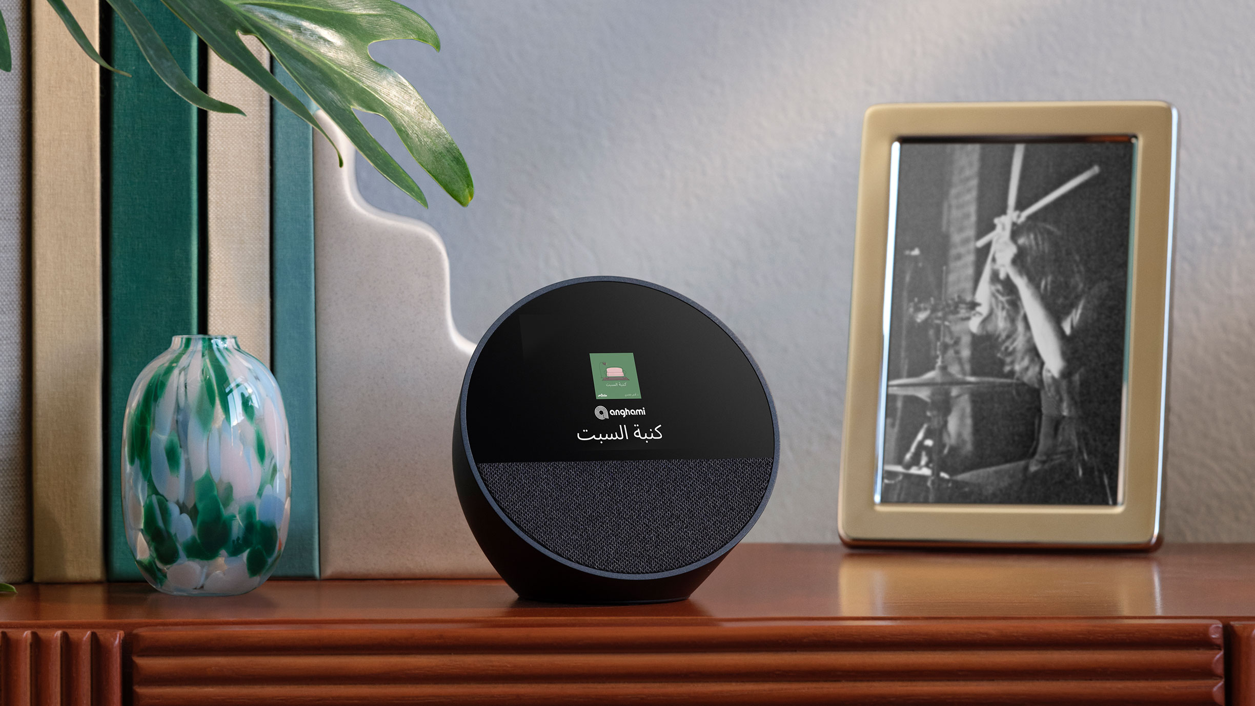 جهاز Echo Spot فوق طاولة مدخل بجانب صورة مؤطرة ومجموعة كتب.  تعرض الشاشة بحجم 2.8 بوصة الوقت وغلاف كتاب صوتي من Anghami.