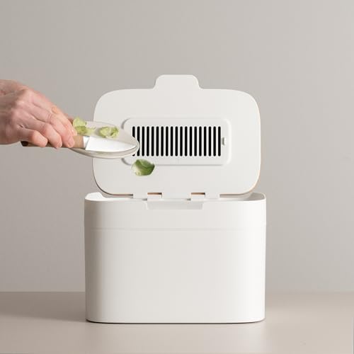 Hand scraping vegetable scraps from a plate into a small white countertop compost bin