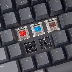 Close-up of a mechanical keyboard showing three different colored Gateron switches (red, blue, brown) removed from their sockets, with the PCB visible, illustrating the hot-swappable feature.