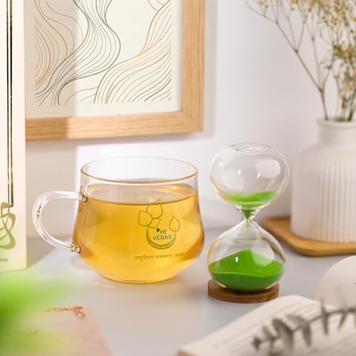 Clear glass mug of tea beside a green sand timer, tea packaging, a framed print, and a white vase in a bright room.