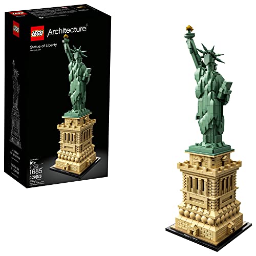 LEGO Architecture Statue of Liberty 21042 displayed on a clean white shelf in a modern American home — the 17-inch tall figure standing on the Liberty Island microscale terrain base with the iconic green-patina copper coloring of the statue itself, the surrounding Liberty Island base showing the star-fortress walkways and the small flagpole, the Architecture line signature display tile naming the set at the front edge of the base — the current Architecture flagship continuously available since 2018