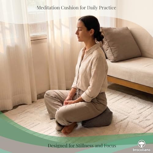 Person sitting upright on a gray zafu-style meditation cushion in a calm home space near a sofa and window curtains.