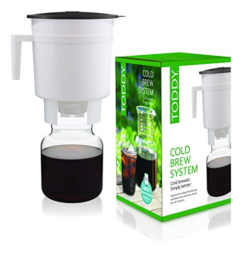 Toddy Cold Brew System on a kitchen counter -- the white plastic brewing container holding a brown coffee-soaked mixture draining through the felt filter stopper into the glass carafe below, the lid and rubber stopper beside it -- the original commercial cold brew system invented in 1964 that makes smooth, low-acid cold brew concentrate at home