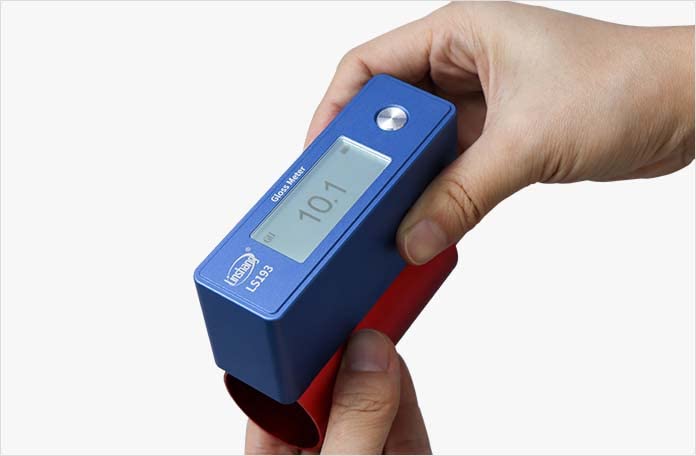Hand holding the LS193 Gloss Meter to measure gloss on a curved red object, displaying a reading of 10.1 GU.