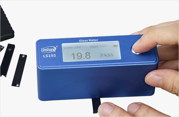 Hand holding the LS193 Gloss Meter to measure gloss on a small black rectangular sample, displaying a reading of 19.8 GU and 'PASS'.