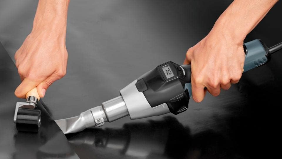 Hands using the Steinel HG 2620 E Heat Gun to apply heat to a black material, with a roller tool.