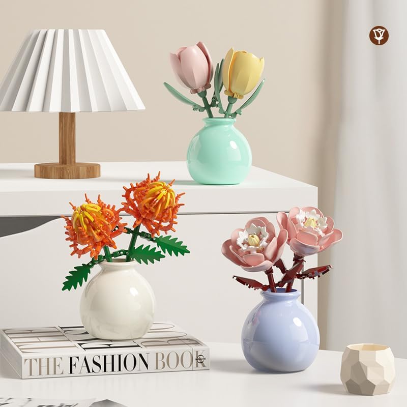 Three assembled mini building block flowers in vases on a white desk: pink and yellow tulips, orange chrysanthemums, and pink peonies.