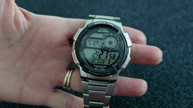 Depan view of a Casio AE1000W digital watch with a silver-tone case and black resin band, displaying world time and a map.
