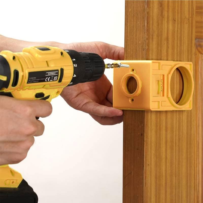 A person's hands holding a yellow TOLSEN positioning frame against a wooden door, securing it with a screw using a cordless drill.