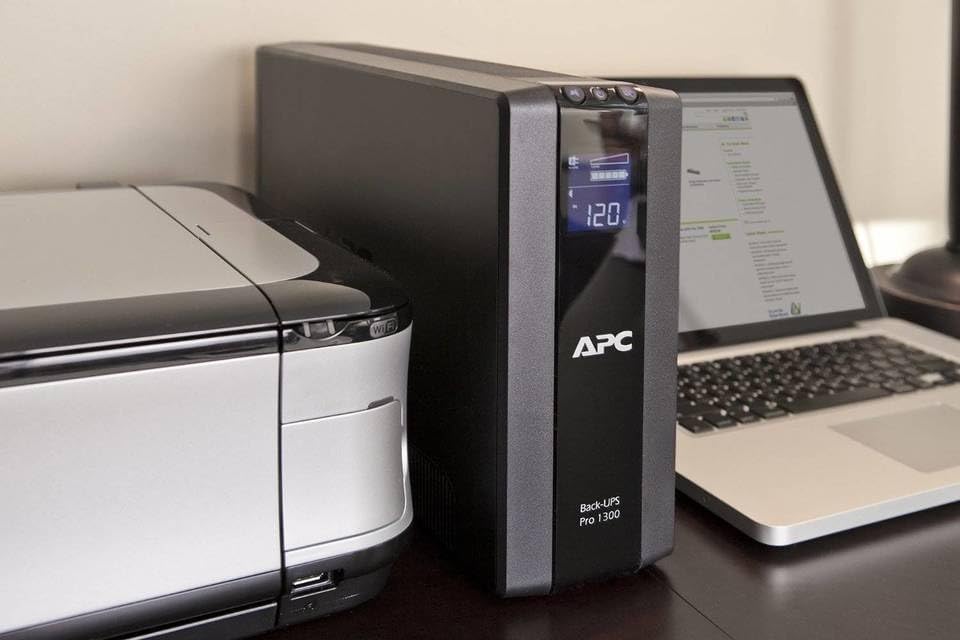APC Back-UPS Pro 1500VA-enhed placeret ved siden af en printer og bærbar computer, hvilket demonstrerer dens kompakte størrelse i en typisk kontoropsætning.