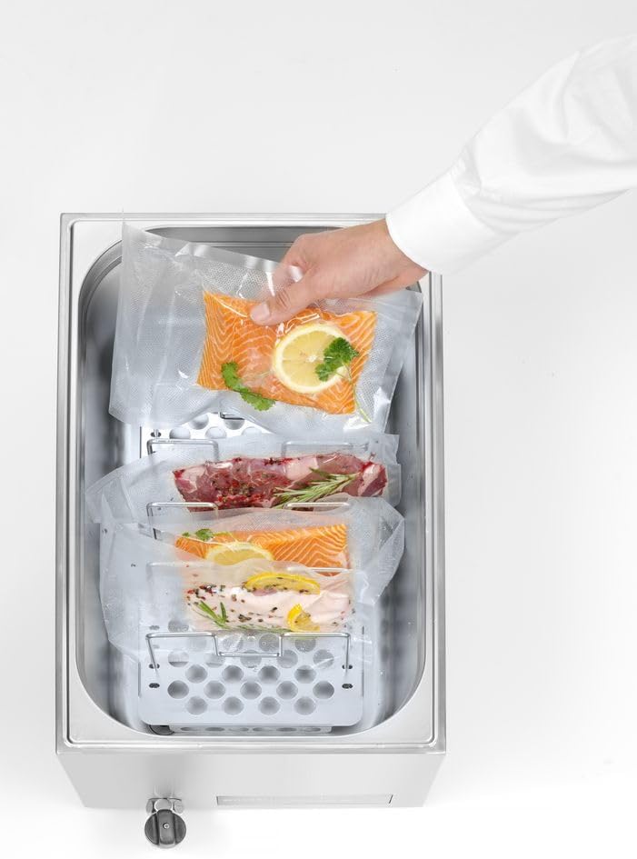 A hand placing a vacuum-sealed salmon fillet into the sous vide cooker, which already contains other food items.