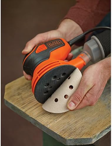 A hand attaching a red 5-inch sanding disc to the base of an orange and black BLACK+DECKER random orbit sander.