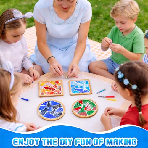 Adult guiding four children as they place colorful mosaic tiles on round wooden coaster projects while sitting on a picnic blanket.