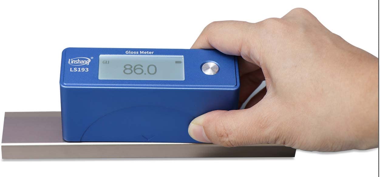 Hand holding the LS193 Gloss Meter to measure gloss on a flat metallic surface, displaying a reading of 86.0 GU.
