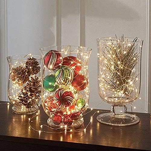 Three clear glass vases on a wooden surface, two filled with Christmas ornaments and fairy lights, and one with fairy lights and pine needles.