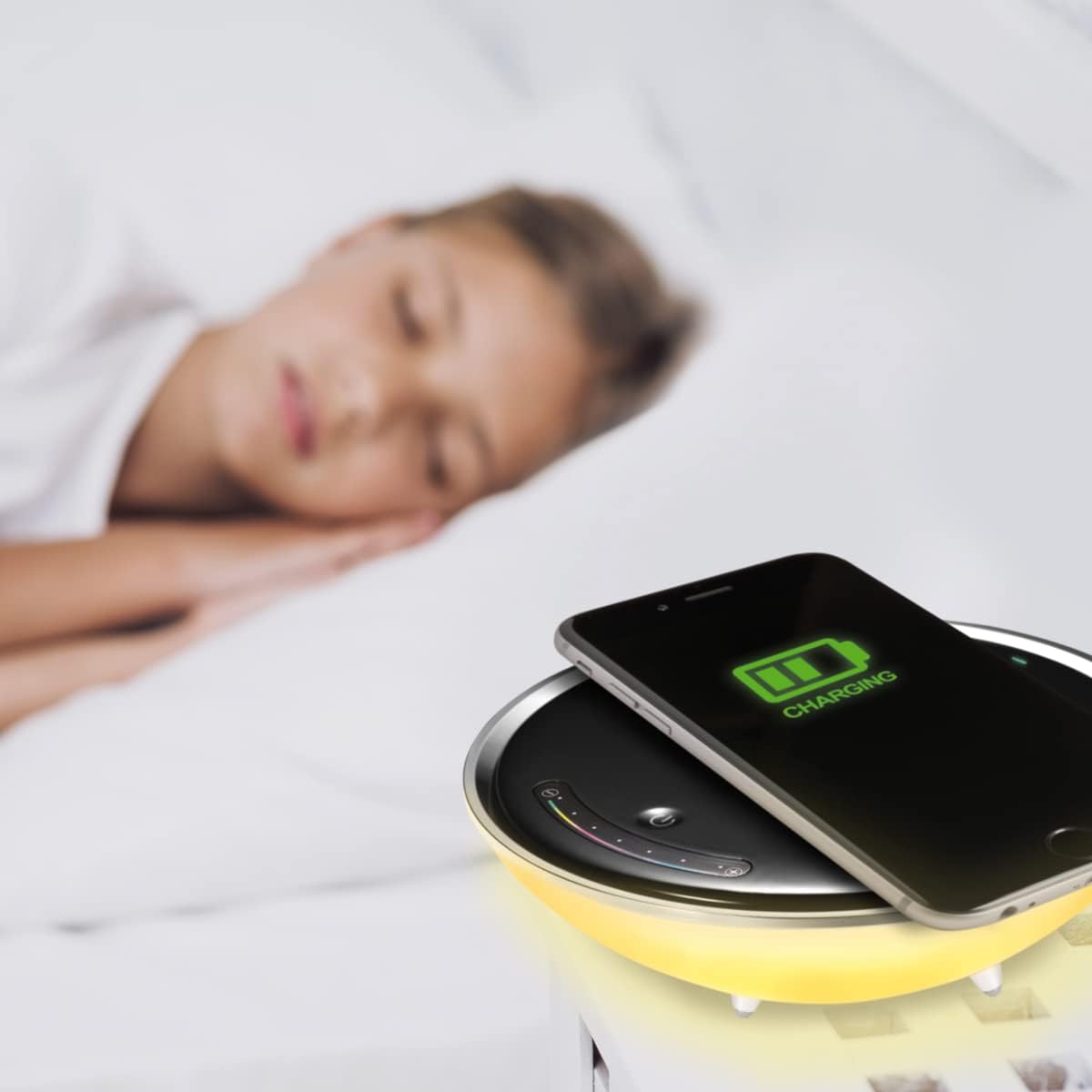 A smartphone charging wirelessly on the PLATINET PDLU1 lamp, with a green charging icon visible on the phone screen. A person is sleeping in the background.