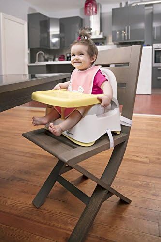 A baby happily seated in the Badabulle B009009 Booster Seat with the yellow tray, attached to a dining chair.