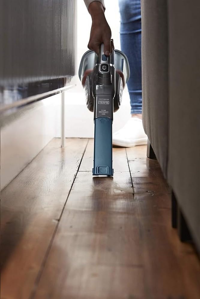 A person using the BLACK+DECKER handheld vacuum to clean dust and debris from under a piece of furniture.