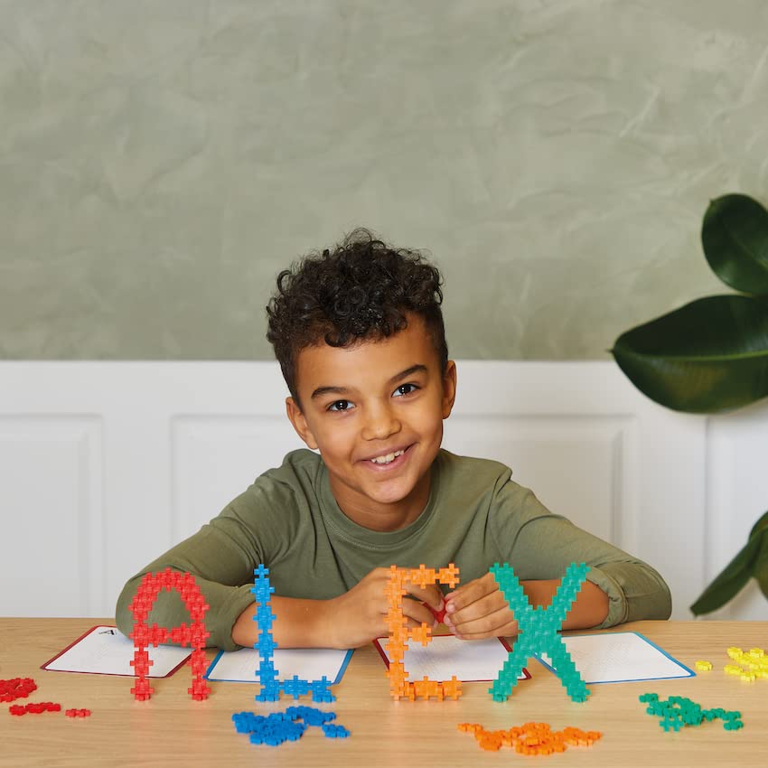 A child smiling while sitting at a table with various letters built from PLUS PLUS pieces