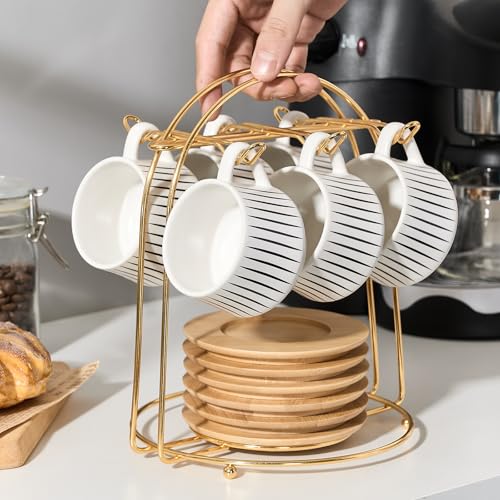 Six white striped ceramic espresso cups hanging from a gold metal holder above stacked wooden-colored saucers beside a coffee machine.